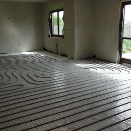 Usługi Ogólnobudowlane Daniel Wyrzykowski - Rozłożone rury ogrzewania podłogowego na siatce w surowym wnętrzu z oknami i nieotynkowanymi ścianami.